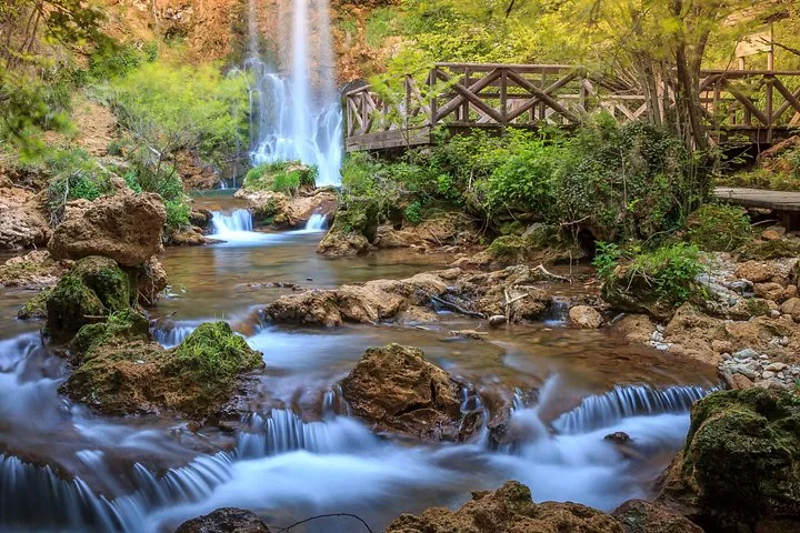 Ein wunderschöner Wasserfall stürzt über Felsen in einen flachen Bach neben einer Holzbrücke.